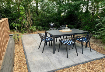 Buiten eethoek bij een lodge, zwart tuinmeubilair op betegeld terras, omringd door groene bomen.