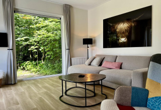 Woonkamer in chalet met groot raam en tuinzicht, moderne bank, kunst aan de muur en ronde salontafel.