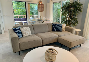 Moderne chaletstue i Holland med lys sofa, blå puder, stort vindue og terrasse med siddepladser.