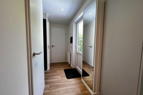 Lichte, moderne inkomhal in Nederlands chalet, met houten vloer, grote spiegel en witte deuren.