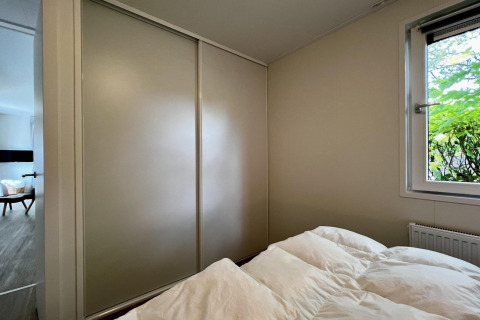 Chambre dans un chalet néerlandais, armoire, fenêtre donnant sur le jardin et lit avec couette blanche.