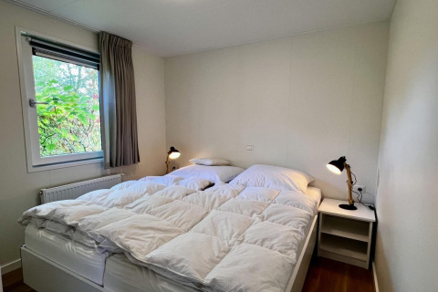 Dormitorio con cama doble, ropa de cama blanca, ventana con cortinas grises y mesitas en chalet holandés.