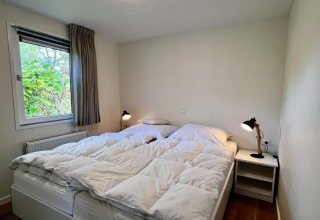 Dormitorio con cama doble, ropa de cama blanca, ventana con cortinas grises y mesitas en chalet holandés.