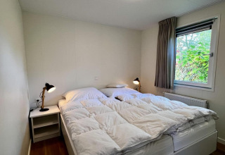 Slaapkamer met tweepersoonsbed, witte dekbedden, nachtkastjes met lampen en raam met tuinzicht.
