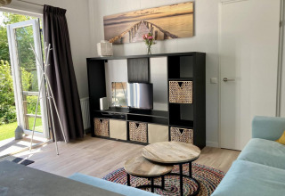 Moderne chalet woonkamer in Nederland met open tuindeur, kast, tafels, zetel en tuinzicht.