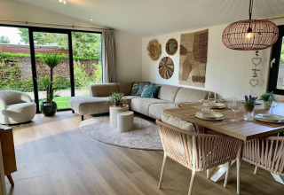 Moderne gezellige woonkamer in een Nederlands chalet met grote ramen, eethoek, hoekbank en planten.