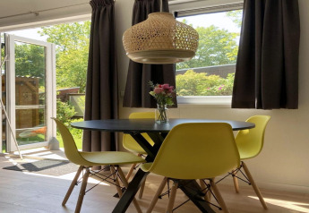 Lyst chaletinteriør med gule stole, sort bord, hængende lampe, og åben dør til grøn have.