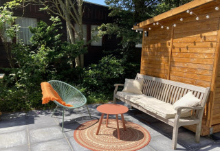 Terraza exterior de chalet moderno en Países Bajos con banco, silla, mesa pequeña y jardín arbolado.