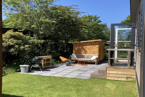 Zonnige tuin bij modern chalet met terras, barbecue en zitbank, omgeven door groen in Nederland.