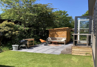 Zonnige tuin bij modern chalet met terras, barbecue en zitbank, omgeven door groen in Nederland.
