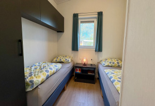Twee eenpersoonsbedden met vrolijk beddengoed, nachtkastje en raam in kamer dichtbij Sint Maartenszee Beach.