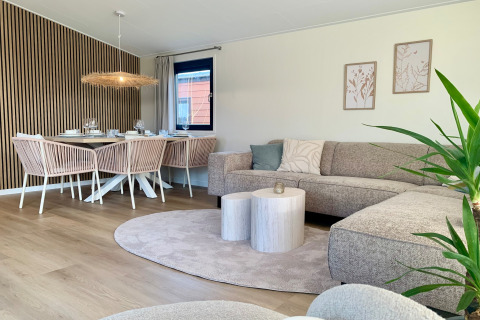 Moderne, lichte leefruimte in strandchalet met eettafel, comfortabele zetel, planten en houten details.