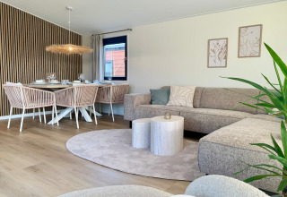 Moderne, lichte leefruimte in strandchalet met eettafel, comfortabele zetel, planten en houten details.