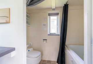 Baño en una tiny house en los Países Bajos con inodoro, bañera, ventana y paredes claras.