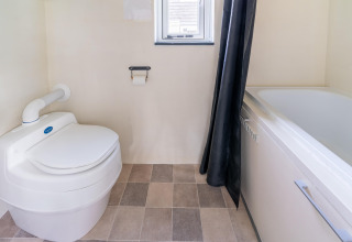 Baño en una tiny house en Países Bajos, con inodoro moderno, bañera, ventana pequeña y suelo de baldosas.