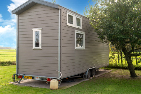 Grijs tiny house op wielen geparkeerd naast een boom en veld onder blauwe lucht in Nederland.