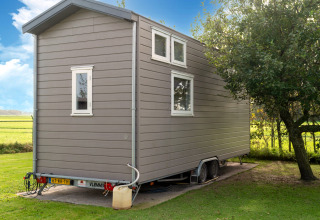 Grijs tiny house op wielen geparkeerd naast een boom en veld onder blauwe lucht in Nederland.