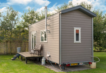 Klein grijs tiny house met terras en stoelen, gelegen op gras in Nederland onder een blauwe lucht.
