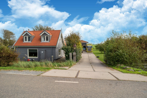 Casa pequeña con tejado rojo y fachada gris, entrada y jardín verde bajo cielo azul en los Países Bajos.