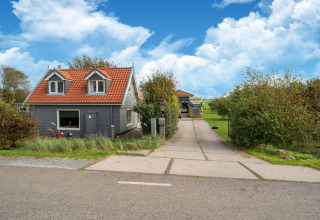 Casa pequeña con tejado rojo y fachada gris, entrada y jardín verde bajo cielo azul en los Países Bajos.