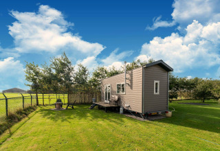 Tiny house op wielen in Nederland, gelegen op gras met een zithoek en omrand door bomen en blauwe hemel.