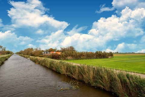 Vlaams platteland met een kanaal, groene velden en tiny house onder een blauwe lucht met wolken.