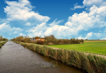 Vlaams platteland met een kanaal, groene velden en tiny house onder een blauwe lucht met wolken.
