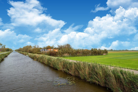 Paisaje holandés con canal, campos verdes y una tiny house bajo un cielo azul con nubes esponjosas.