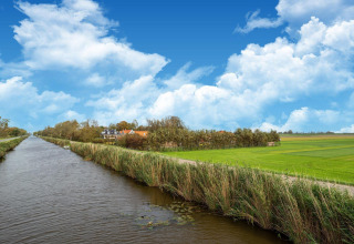 Paisaje holandés con canal, campos verdes y una tiny house bajo un cielo azul con nubes esponjosas.
