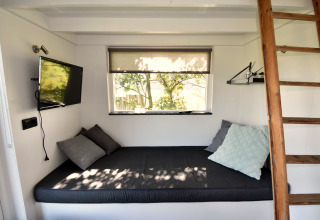 Gezellig interieur van tiny house in Nederland met bed, kussens, tv, raam en houten ladder.