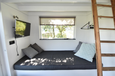 Interior luminoso de tiny house en Países Bajos con cama, cojines, TV y escalera de madera junto a ventana.