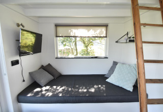 Interior luminoso de tiny house en Países Bajos con cama, cojines, TV y escalera de madera junto a ventana.