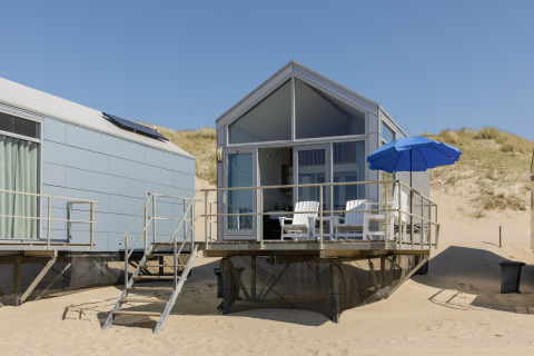 Casa sulla spiaggia moderna a Resort Beach Houses Julianadorp 1 con patio, sedie e ombrellone blu sulla sabbia.