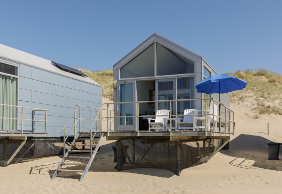 Casa sulla spiaggia moderna a Resort Beach Houses Julianadorp 1 con patio, sedie e ombrellone blu sulla sabbia.