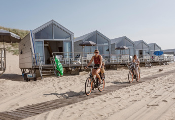 Twee vrouwen fietsen langs strandhuisjes bij Resort Beach Houses Julianadorp in Nederland op een zonnige dag.