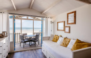 Zonnige woonkamer in strandhuis met zeezicht, witte bank, gele kussens en zitje op het terras.