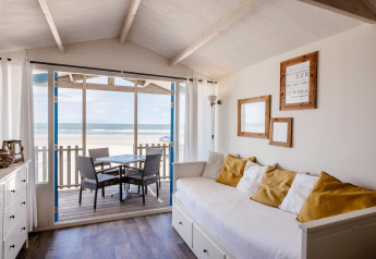Zonnige woonkamer in strandhuis met zeezicht, witte bank, gele kussens en zitje op het terras.