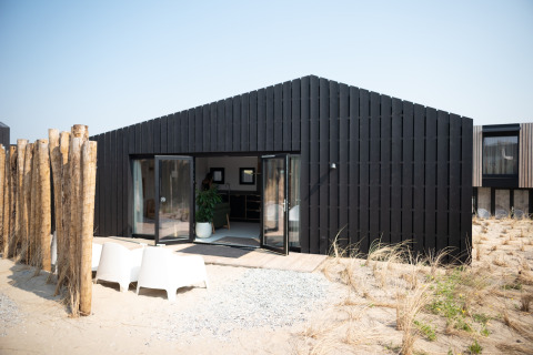 Casa de playa moderna y negra en Sea Lodges Zandvoort 3 con entorno arenoso y sillas exteriores.