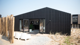 Casa de playa moderna y negra en Sea Lodges Zandvoort 3 con entorno arenoso y sillas exteriores.