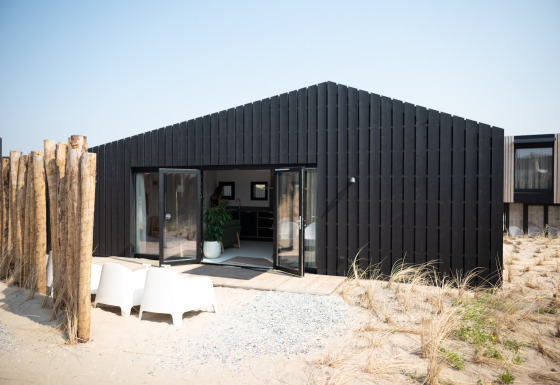 Casa de playa moderna y negra en Sea Lodges Zandvoort 3 con entorno arenoso y sillas exteriores.