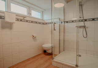 Moderne badeværelse med glasbrusekabine, vægmonteret toilet og hvidt fliseindretning på husbåd.
