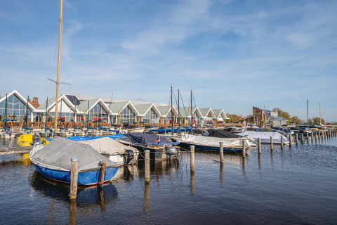 Boten aangemeerd aan een steiger met moderne glampinghuisjes op de achtergrond onder blauwe lucht.
