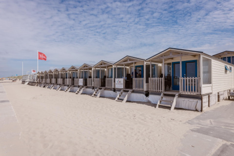Row of beach houses with blue doors lined up on the sand under a partly cloudy sky on a calm day.
