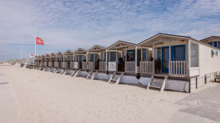 Rij strandhuisjes met blauwe deuren op het zand onder een licht bewolkte hemel op een rustige dag.