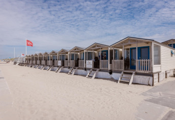 Rij strandhuisjes met blauwe deuren op het zand onder een licht bewolkte hemel op een rustige dag.