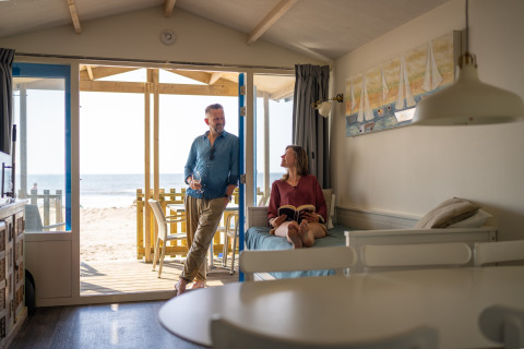 A couple relaxes in a beach house, with the man standing in the doorway and the woman reading on the sofa.