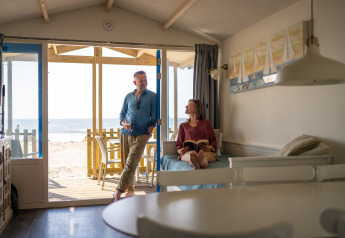 Een stel ontspant in een strandhuisje; de man staat bij de deur en de vrouw leest op de bank.