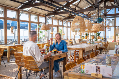 Two people enjoying a meal together in a bright beach house restaurant with wooden tables and wicker lamps.