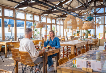 Twee mensen genieten samen van een maaltijd in een licht strandhuisrestaurant met houten tafels en rieten lampen.