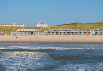 Strandhuisjes en een strandpaviljoen aan de kust met grasduinen op de achtergrond onder een blauwe lucht.